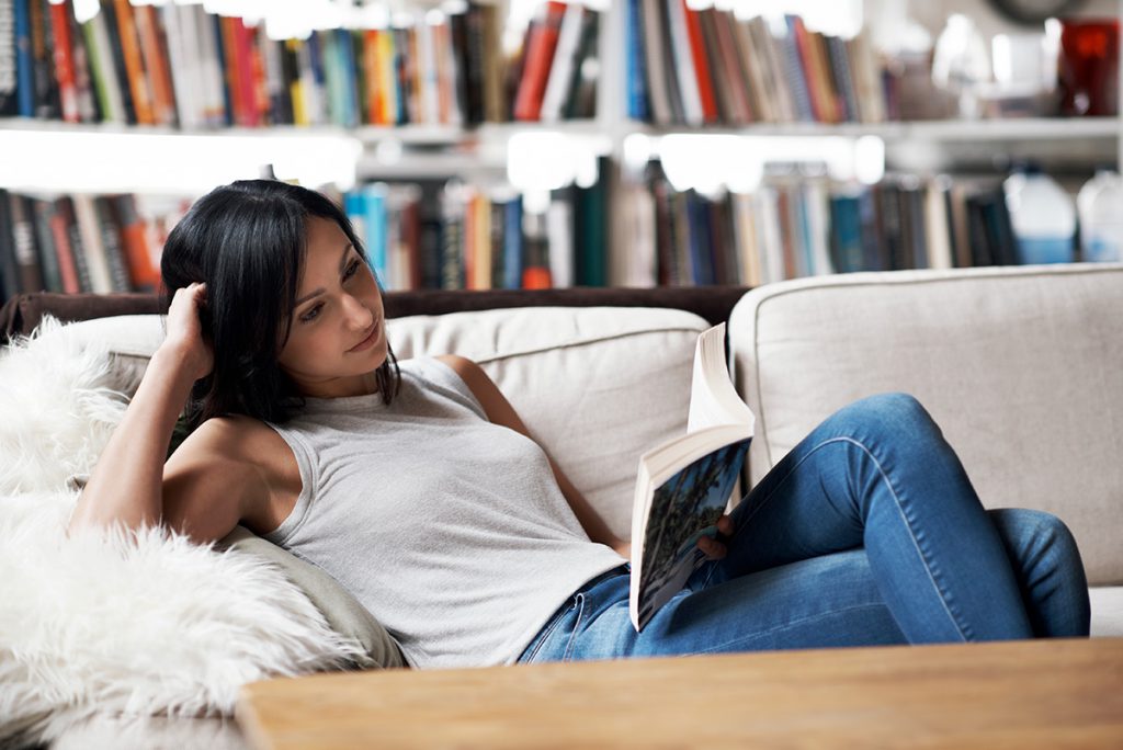 Imagem mostra moça lendo um livro sentada no sofá 